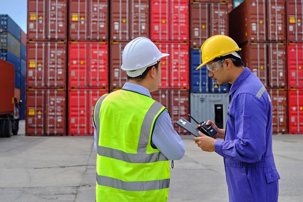 Two male workers work at a logistics terminal with many stacks of containers.